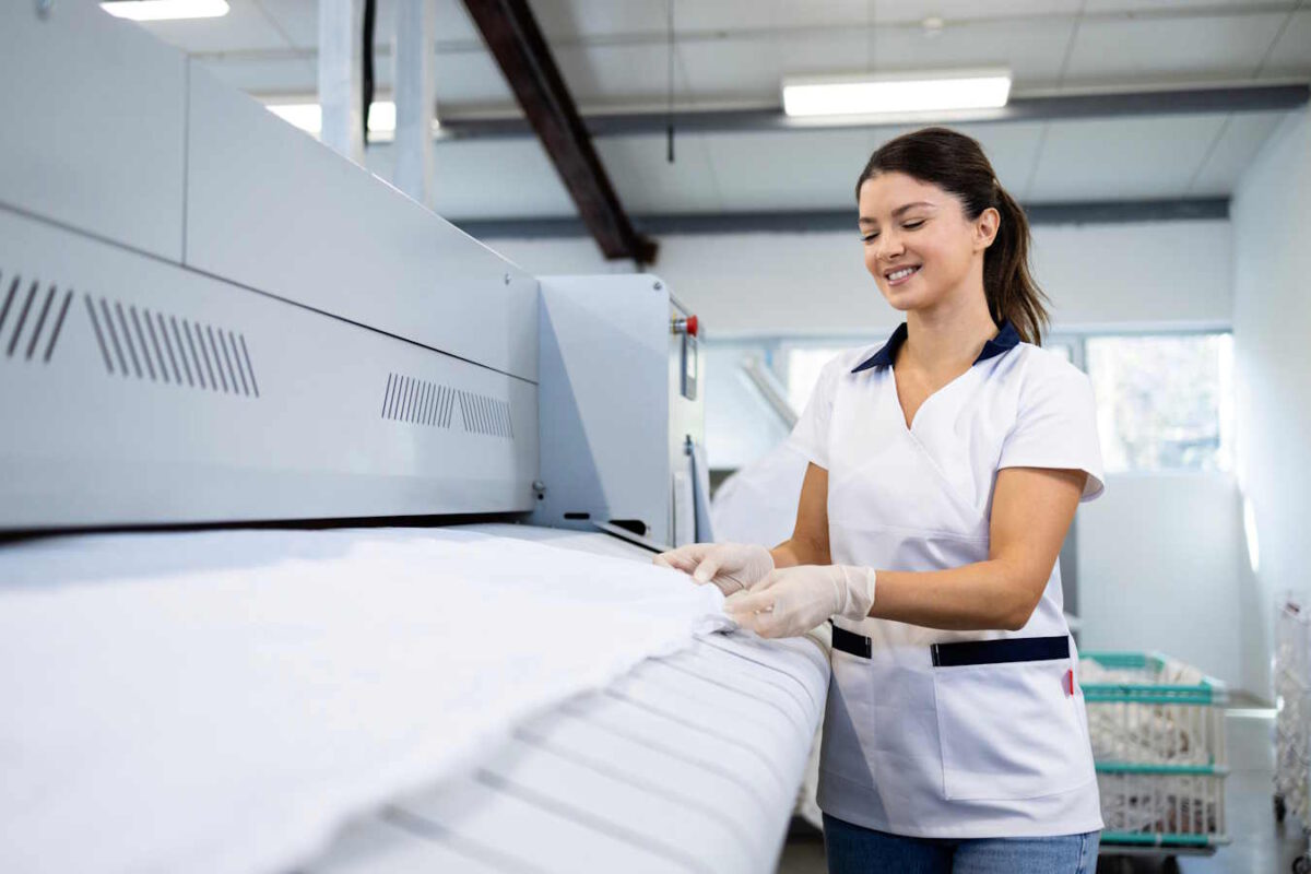 Eine Frau steht vor einer industriellen Bügelmaschine. Sie bereitet Mietwäsche für Hotels auf.